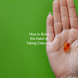 Hand holding two Calocurb capsules against a green background, symbolizing habit formation and daily supplement routine